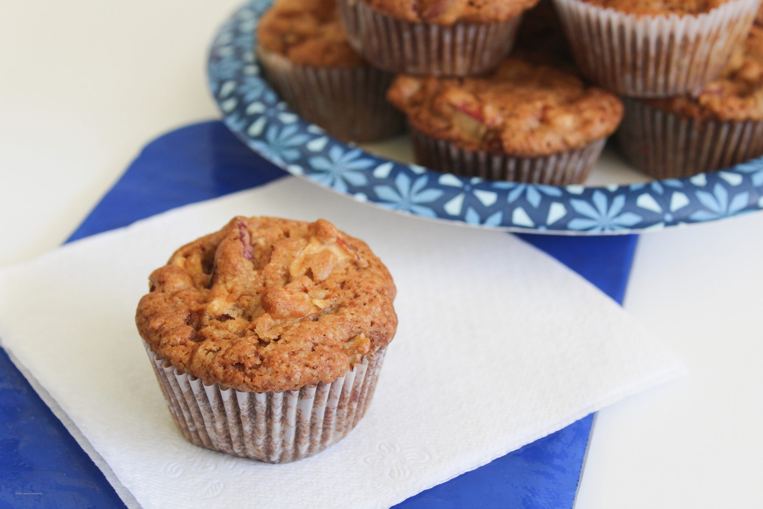 A golden brown apple cake muffin sitting on a napkin with a plate full of other muffins behind it.