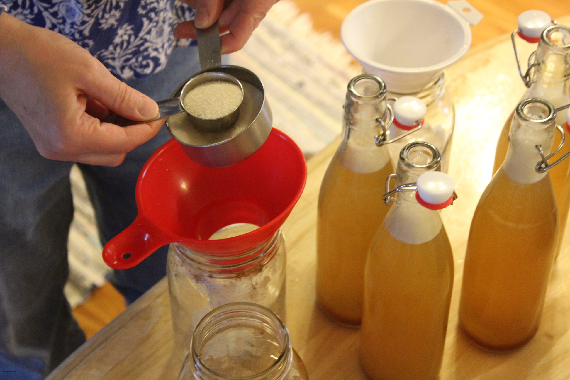 Spooning white sugar into a jar of water kefir with bottles of water kefir to the side.